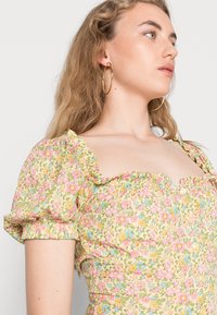 Vestuário floral em amarelo claro com mangas bufantes, apresentando padrões florais em rosa, azul e verde. Decote quadrado com pequenos botões.