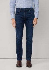 Muškarac nosi uske tamnoplave traperice, svijetloplavu prugastu košulju uvučenu unutra i smeđe kožne čizme, a stoji uz jednostavnu pozadinu.