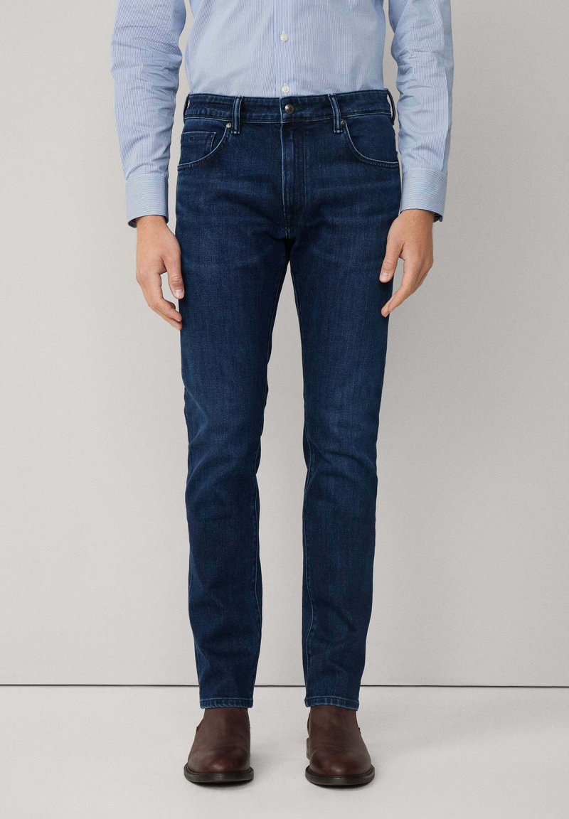 Muškarac nosi uske tamnoplave traperice, svijetloplavu prugastu košulju uvučenu unutra i smeđe kožne čizme, a stoji uz jednostavnu pozadinu.