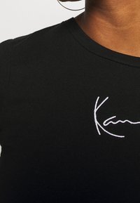 Černé bavlněné tričko s kulatým výstřihem a krátkými rukávy. Na přední straně má bílé vyšívané písmeno "Kaan", hladký povrch.