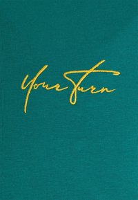 Teal shirt featuring elegant yellow embroidered text that reads “Your Turn,” with a smooth, textured fabric.