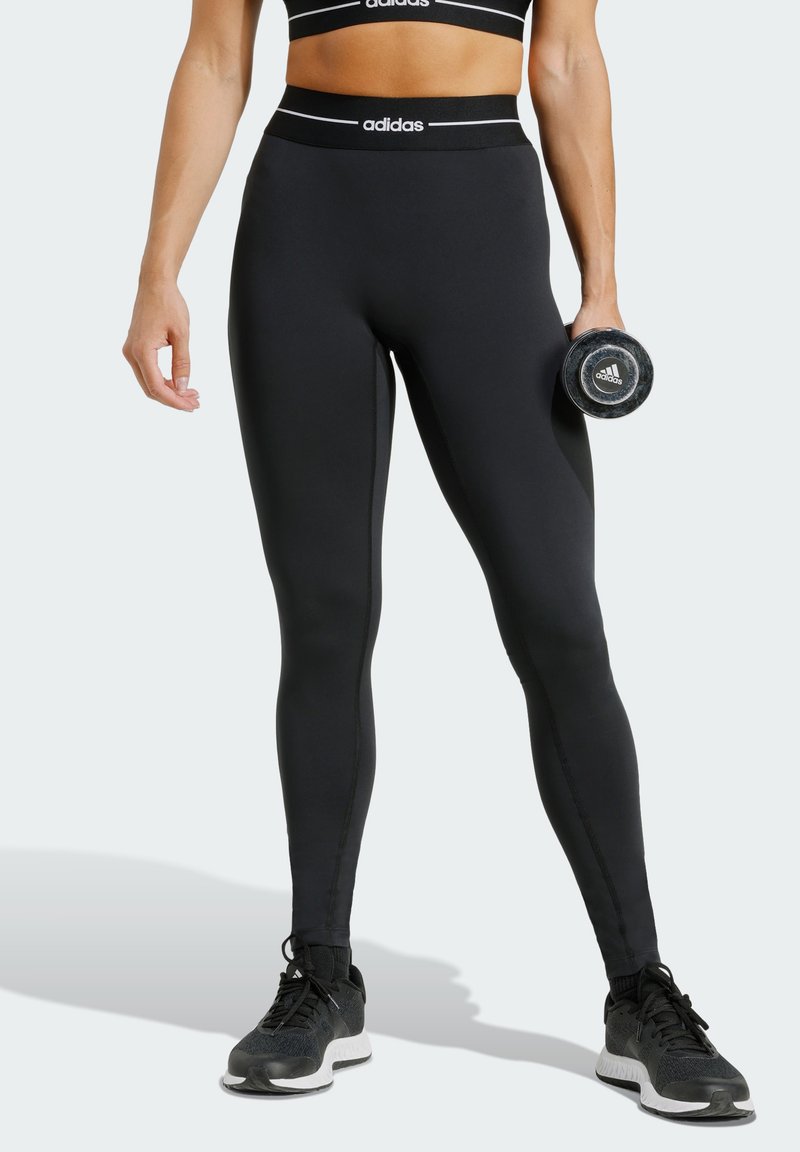 Black leggings with a high waistband featuring white Adidas branding, made of stretchy material, paired with black athletic shoes.