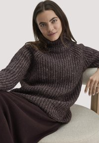 Kuscheliger, dunkelbrauner Strickpullover mit hohem Kragen, geripptem Textur und lässiger Passform, kombiniert mit einem tiefbraunen Stoff darunter.