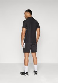 Svart polo-skjorte med korte ermer og hvite sideakenter, kombinert med mørke shorts. Modellen viser atletisk passform og nøytral fargeskjema.