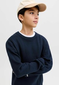 Garçon portant une casquette beige et un pull bleu marine, les bras croisés, regardant sur le côté sur un fond clair uni.