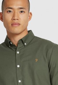 Camicia in cotone verde oliva con bottoni, colletto a punta, bottoni trasparenti e logo arancione ricamato sul lato sinistro del petto.
