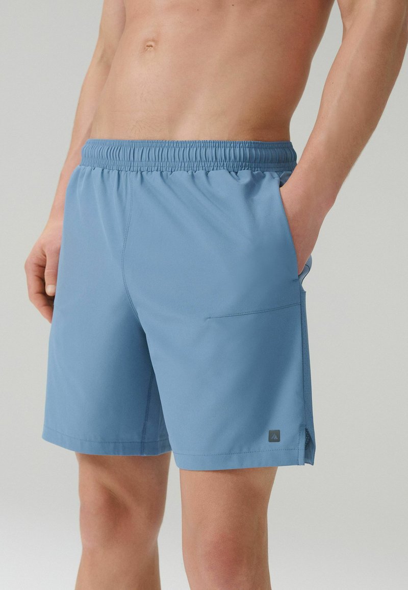 Homme portant un short bleu clair à taille élastique avec poches latérales, debout devant un fond gris clair uni.