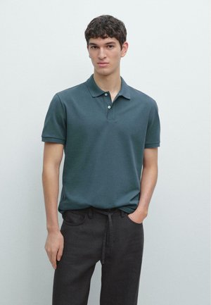 Giovane uomo con capelli corti e ricci che indossa una polo a maniche corte verde petrolio scuro e pantaloni neri, in piedi contro una parete chiara e semplice.