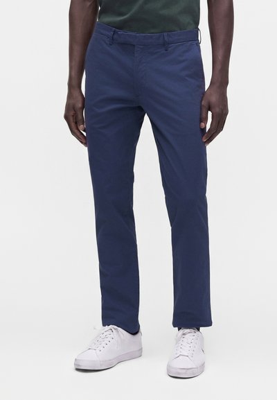 Άνδρας που φοράει σκούρο μπλε παντελόνι στενής εφαρμογής και λευκά αθλητικά με κορδόνια σε απλό φωτεινό φόντο.