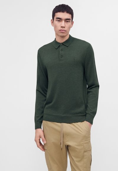 Jeune homme portant un polo vert foncé à manches longues et un pantalon cargo beige, debout avec une main dans la poche.