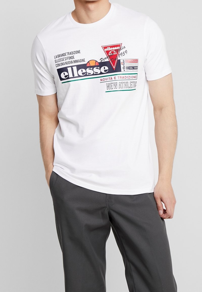 Homme portant un t-shirt blanc à manches courtes avec un texte et un logo colorés de la marque Ellessee, associé à un pantalon gris foncé, debout devant un fond gris.