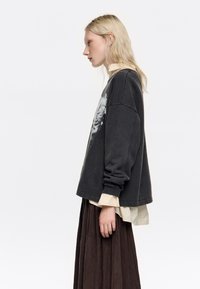 Sudadera de gran tamaño en gris oscuro con estampado gráfico en la parte delantera, superpuesta a una camisa beige clara con cuello y una falda marrón fluida.