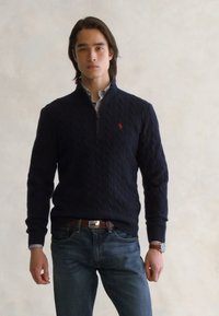 Marine gebreide trui met een kwart-rits halslijn, kabelpatroon en een klein rood logo. Gecombineerd met blauwe jeans en een bruine riem.