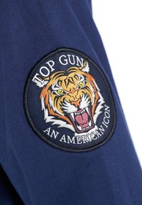 Marinblått tyg med ett broderat märke som föreställer ett vrålande tiger och texten "TOP GUN AN AMERICAN ICON" i vitt och gult.