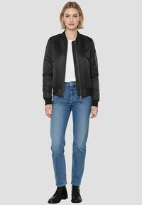 Czarna kurtka bomberka o gładkiej, błyszczącej teksturze, z zamkiem z przodu, ściągaczami na rękawach oraz boczną kieszenią. Połączona z niebieskimi dżinsami i czarnymi botkami za kostkę.