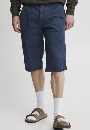 Pantaloni corti in cotone blu navy con una vestibilità rilassata, lunghezza fino al ginocchio e dettagli cuciti. Abbinati a calze bianche e sandali marroni.