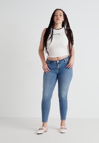 White sleeveless top with black lettering, paired with blue skinny jeans. Model wears white slip-on sandals. Plain background.