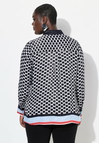 Camisa de patrones en blanco y negro con un diseño geométrico, puños contrastantes con un acento en azul y rojo, textura suave y detalles de botones.