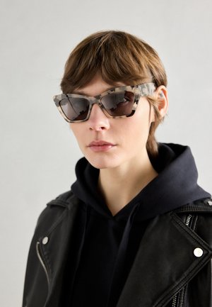 Kaķacīšu saulesbrilles ar bruņurupuču rakstu, tumšiem objektīviem un stūrainu rame, valkātas ar melnu kapuci un ādas jaku.