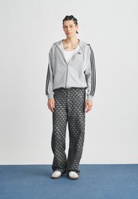 Sweatshirt cinzento com fecho de correr e riscas pretas, combinado com calças largas pretas com padrão de folhas, completo com ténis brancos.