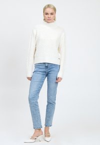 Witte kabelgebreide trui met een hoge kraag, gecombineerd met lichtblauwe jeans en witte muiltjes met hak. De textuur is zacht en gemusterd.