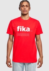 Röd bomull t-shirt med ordet "fika" i fetvita bokstäver och en beskrivning av konceptet nedanför. Rund halsringning och korta ärmar.