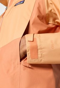 Lätt tvåfärgad jacka i orange och persikotoner. Har en zippered ficka och justerbar mudd med kardborreknäppning. Slät textur.