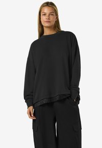 Felpa nera a maniche lunghe con scollatura rotonda, caratterizzata da un bordo di pizzo all'orlo. Abbinata a pantaloni cargo neri larghi.