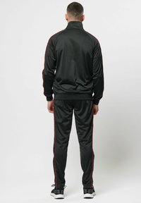 Zwart trainingspak met een ritsjack met hoge kraag en bijpassende broek, beide voorzien van rode zijstrepen en gemaakt van een gladde stof.