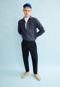 Marineblauw overhemd met knopen, gedragen over een witte colbert, gecombineerd met zwarte broek en tan instapschoenen, afgemaakt met een blauwe pet.