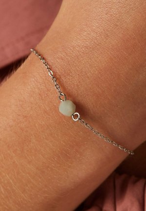 Zilveren kettingarmband met een enkele lichtgroene, ronde kraal. Nette textuur, minimalistisch ontwerp, gedragen om een gebruinde pols.