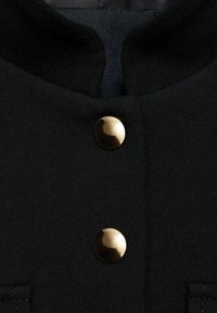 Black fabric garment with a high collar and gold-toned metal button accent. Features smooth texture and clean, tailored lines.