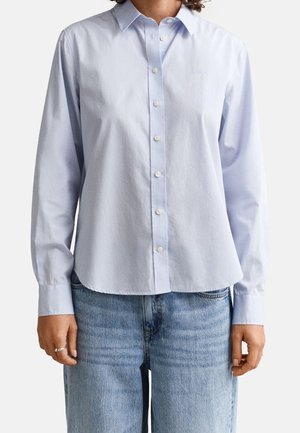 Personne portant une chemise bleu clair à boutons avec manches longues et un jean bleu moyen, debout devant un fond uni.