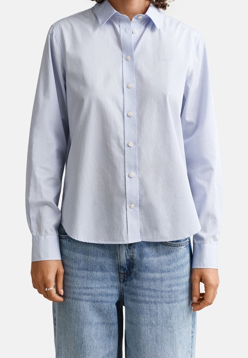 Personne portant une chemise bleu clair à boutons avec manches longues et un jean bleu moyen, debout devant un fond uni.