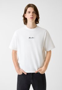 Biała koszulka z krótkim rękawem i okrągłym dekoltem, z czarnym haftowanym logo "Guess Jeans", w zestawieniu z ciemnymi dżinsami. Miękka bawełniana tkanina.