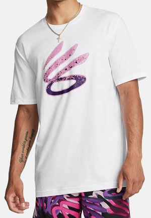 Man draagt een wit T-shirt met een grote roze en paarse abstracte afbeelding en meerkleurige korte broek met paarse en roze patronen.