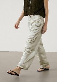 Personne marchant en portant un pantalon en satin beige, des tongs noires et un haut court sans manches sombre avec une fermeture éclair à l'avant.