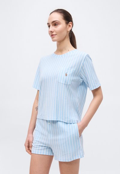 Νέα γυναίκα που φοράει βαμβακερό μπλουζάκι με κάθετες ρίγες σε ανοιχτό μπλε και λευκό, συνοδευόμενο από σορτσάκι στο ίδιο σχέδιο, όρθια με το ένα της χέρι στην τσέπη.