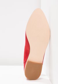 Semelle d'une chaussure rouge en daim à bout pointu avec une semelle antidérapante beige et un talon marron, taille 37, posée à angle droit contre des murs blancs.
