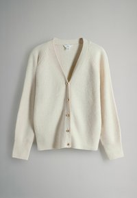 Cremefarvet ribstrikket cardigan med dyb V-udskæring, otte guld-tone knapper og lange ærmer. Blød tekstur, let oversized pasform.