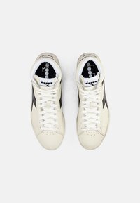 Baskets beige avec des accents noirs et des lacets blancs. Tige en cuir lisse, boîte à orteils perforée, col de cheville rembourré, logo de la marque visible.