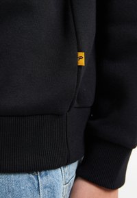 Sudadera negra de tela texturizada, con puños y dobladillo acanalados, que presenta una pequeña etiqueta amarilla en el lateral. Llevada sobre unos vaqueros de color azul claro.