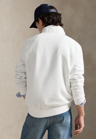 Balta džemperis ar augstu apkakli un uzlocītiem aprocēm, kopā ar zilām džinsa biksēm. Audums šķiet mīksts ar gludu tekstūru.