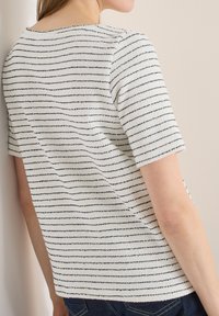 Personne portant une chemise blanche à manches courtes avec de fines rayures horizontales noires, debout contre un mur de couleur claire.