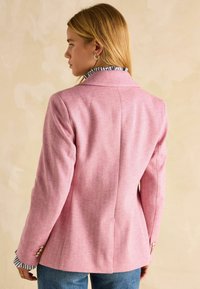 Roosa herringbone ülikond, millel on struktureeritud lõige, silmapaistvad kraed ja kuldse nööbiga aktsendid varrukatel, kantud triibulise särgi peale.