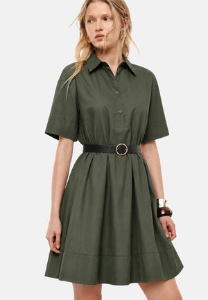 Donna che indossa un vestito verde oliva a maniche corte con colletto e cintura nera con fibbia rotonda dorata. Ha i capelli biondi e lunghi e indossa dei braccialetti.