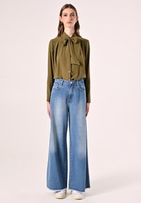 Olivegrüne Bluse mit Schleife am Kragen, Knopfdetails, kombiniert mit weit geschnittenen hellblauen Denim-Jeans und weißen Canvas-Schuhen.