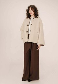 Giacca beige in peluches con una forma ampia, caratterizzata da due bottoni scuri e tasche laterali, abbinata a pantaloni larghi marroni.