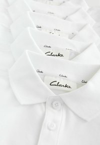 Fem vita polo-shirtar staplade, som visar kragar och knappar, med synliga "Clarks"-etiketter inuti varje skjorta.