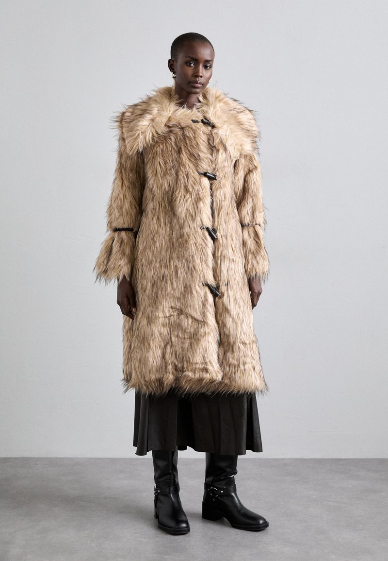 Faux fur jas in lichtbeige met een volumineuze kraag, sluitingen met toggles en lange mouwen. Draagt over een donkere plissé rok en zwarte laarzen.